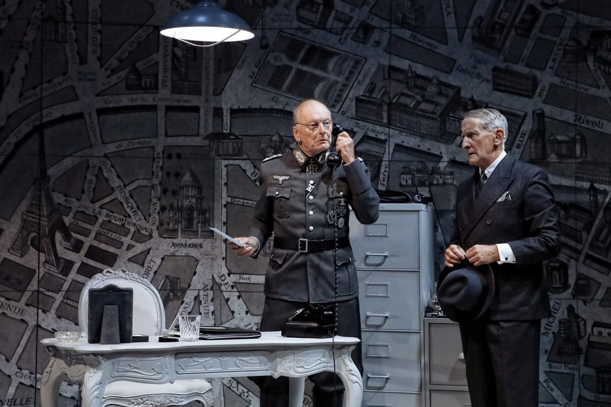 Two older men stand in an office. One wears a military uniform and speaks on a vintage telephone, while the other, in a suit holding a hat, looks on. The backdrop is a large, detailed map. A lamp hangs above a white desk.