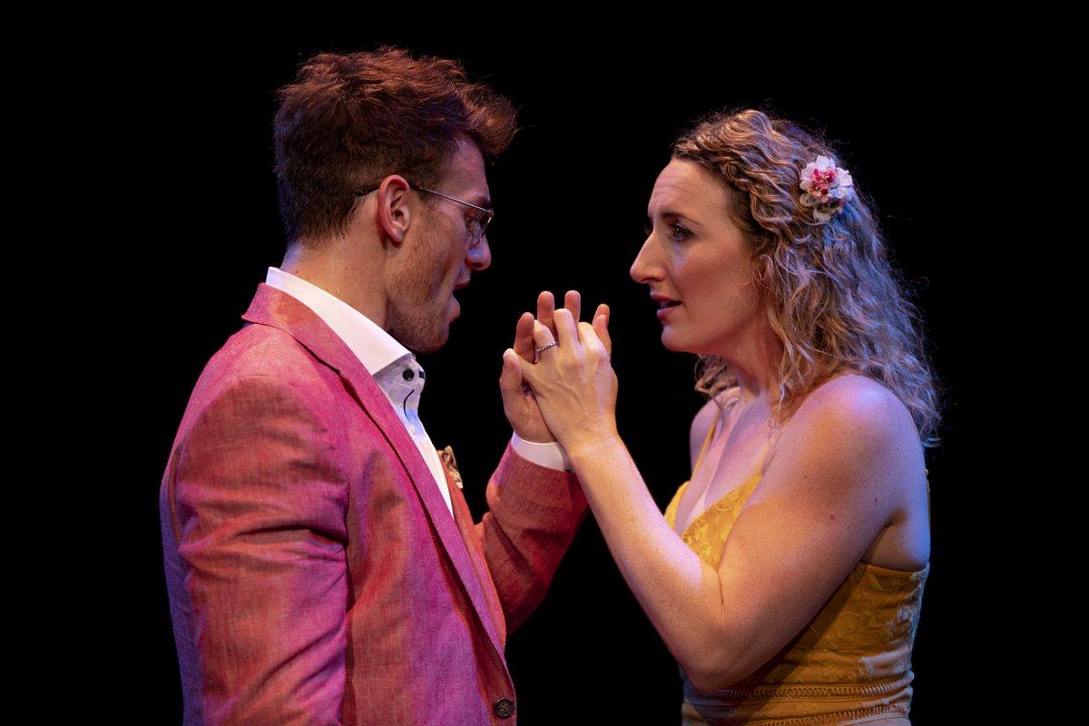 A man in a pink suit and glasses faces a woman in a yellow dress with a flower in her hair. They hold hands and look intently at each other against a dark background.