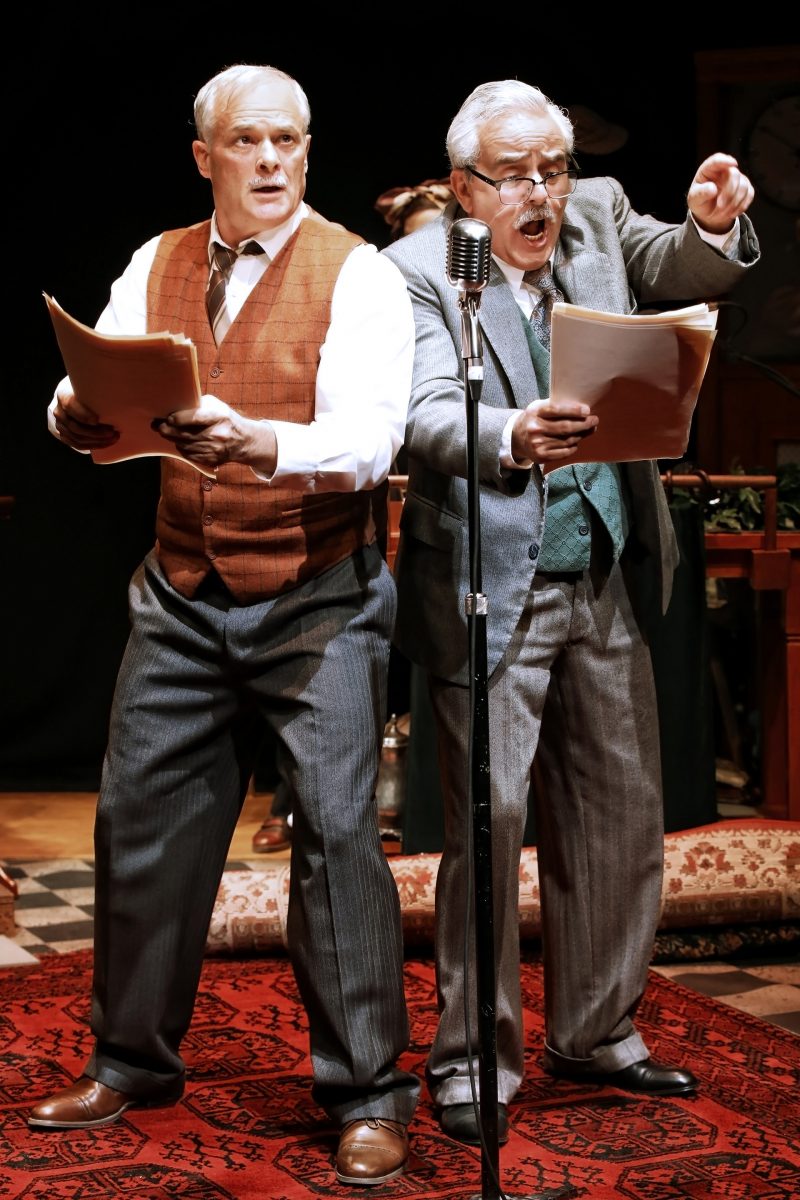 Two older men in vintage clothing stand on a patterned red carpet, holding scripts and performing at an old-fashioned microphone; one looks alert, the other points and speaks passionately.