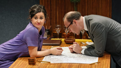 A woman in a purple dress sits at a wooden table, looking sceptically at the camera while a man in a suit leans over a map on the table. The setting appears to be an office or study with glasses and papers in the background.
