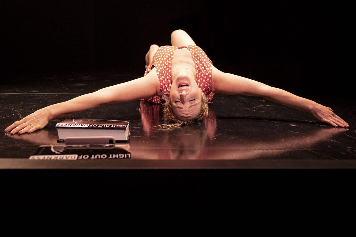 A woman in a red polka dot dress lies on her back on a shiny black surface, arms outstretched, head tilted back, and face expressive. A box labelled LIGHT OUT OF DARKNESS sits nearby.