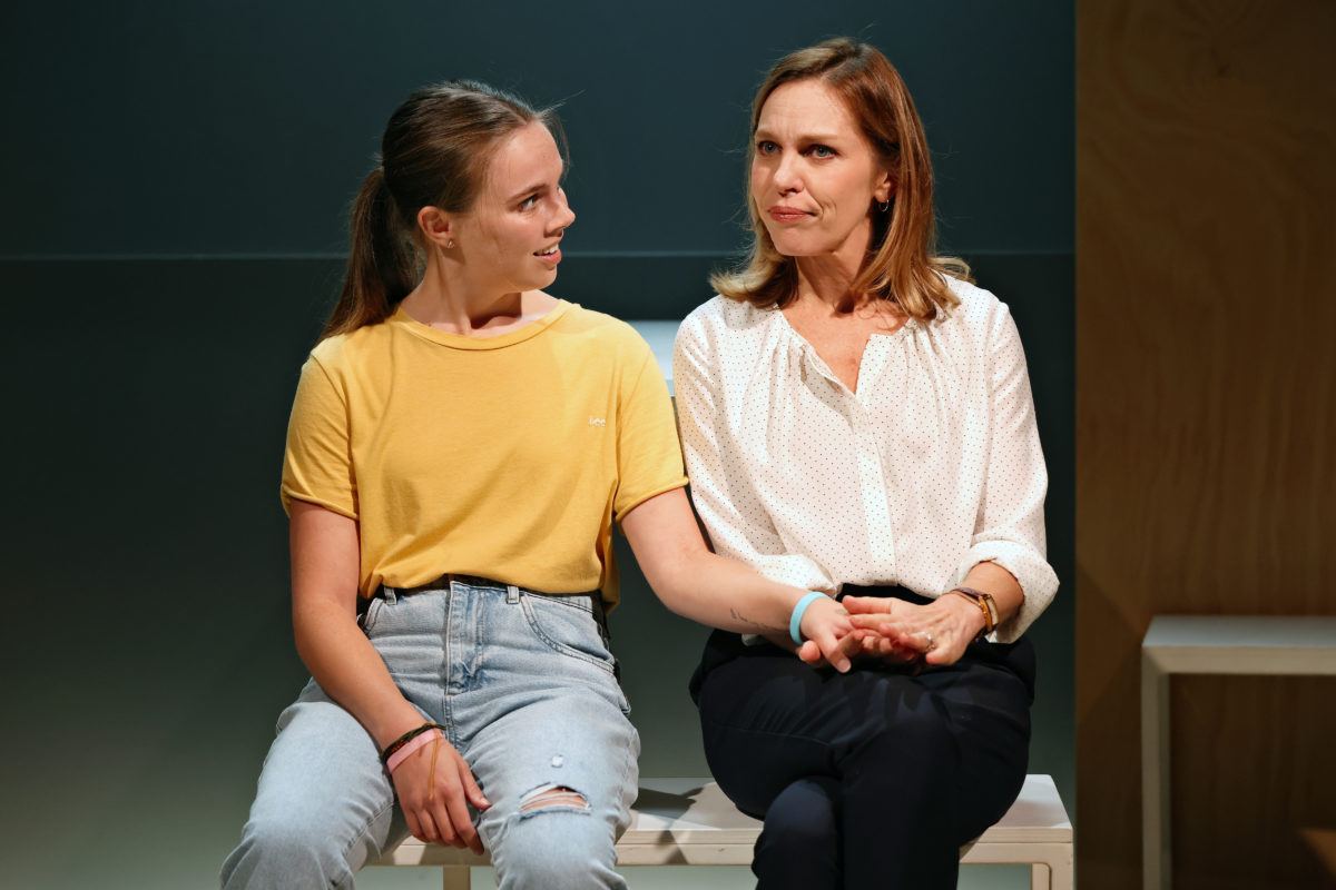 A young woman in a yellow T-shirt and jeans sits beside an older woman in a white blouse and dark trousers. The young woman holds the older woman’s arm, and both look at each other with thoughtful expressions.