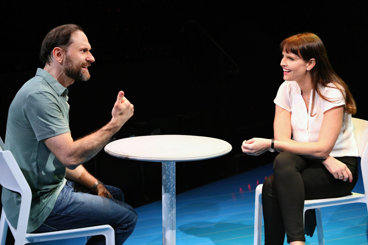 A man and a woman sit opposite each other at a small round table, engaged in a lively conversation. The man gestures with his hand, while the woman smiles attentively. Both are seated on white chairs against a dark background.