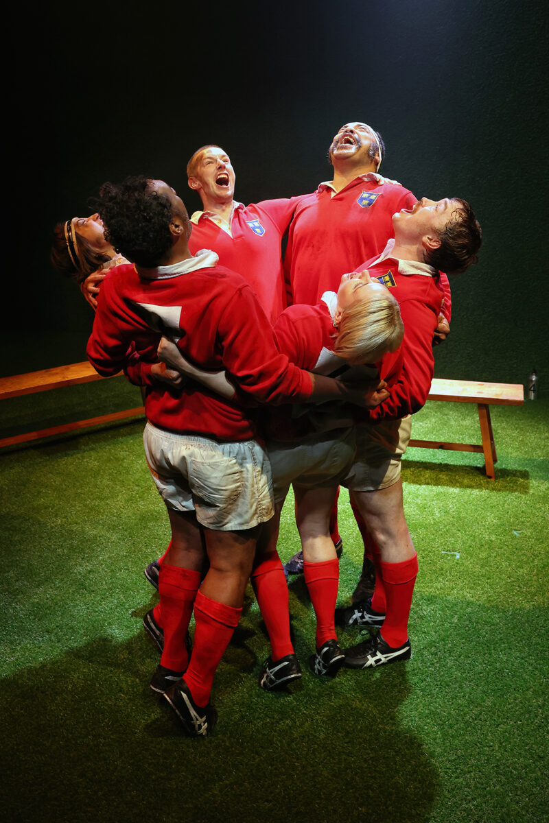 Six rugby players in red jerseys stand in a tight huddle on a green pitch, arms round each other, laughing joyfully and looking upwards. A wooden bench is visible in the background.