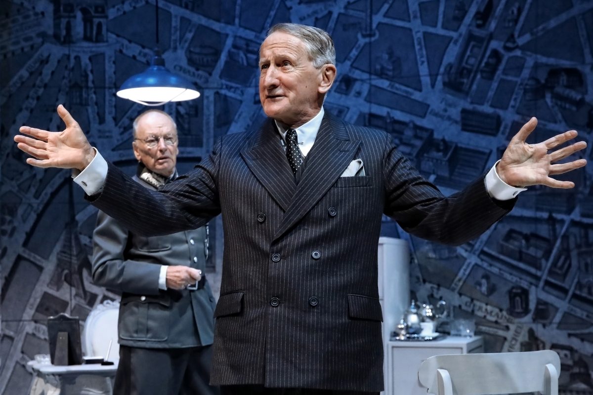 Two older men in suits stand in a room with a blue map-patterned wall; one man gestures widely whilst speaking, and the other stands in the background watching. A desk and tea set are visible nearby.