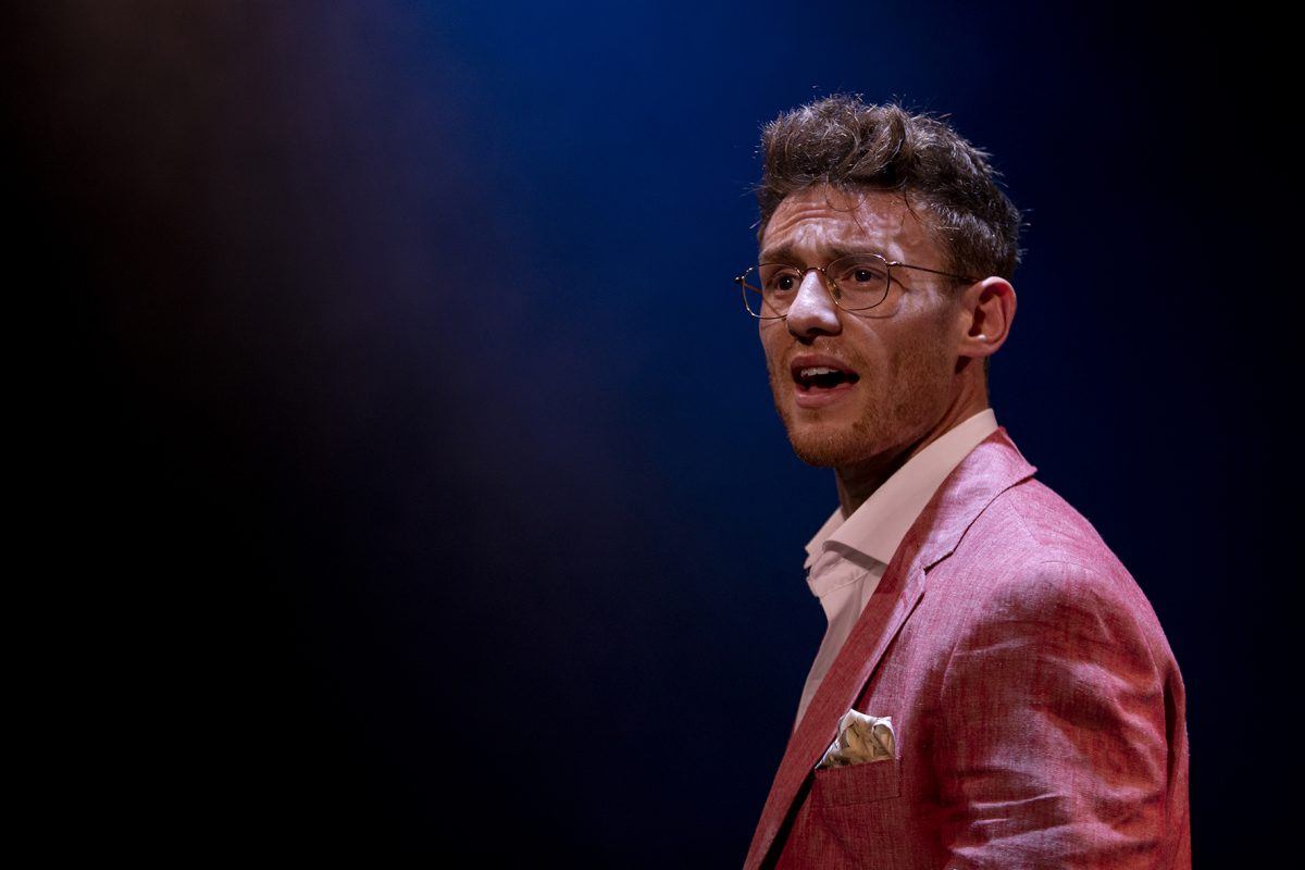 A man in a pink suit with glasses stands under dramatic lighting, looking towards the camera with a serious expression. The background is dark, with a subtle blue and brown gradient.