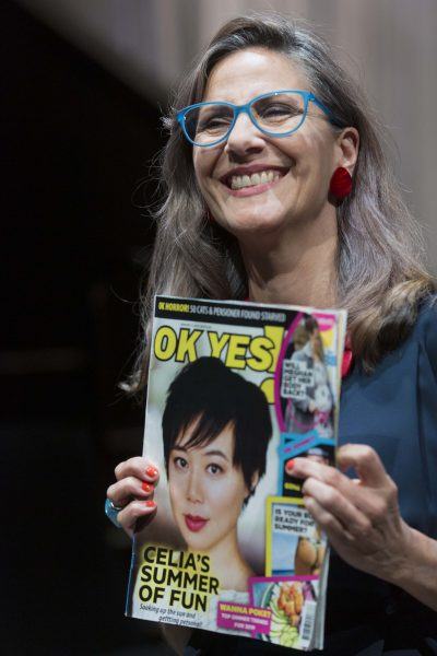 A smiling woman with grey hair and blue glasses holds up a magazine called OK YES! featuring a cover story titled Celia's Summer of Fun with a photo of another woman on the cover.
