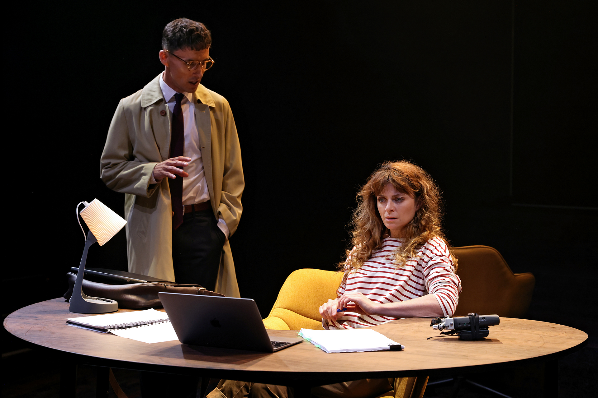 A man in a trench coat and glasses stands by a round desk, speaking to a seated woman in a striped shirt. The desk holds a laptop, papers, a lamp, and a recorder. The scene is dimly lit, suggesting a serious conversation.