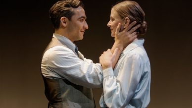 A man gently holds a woman's face with both hands, gazing at her lovingly. Both wear vintage-style clothing, and stand close together against a plain, brown background.