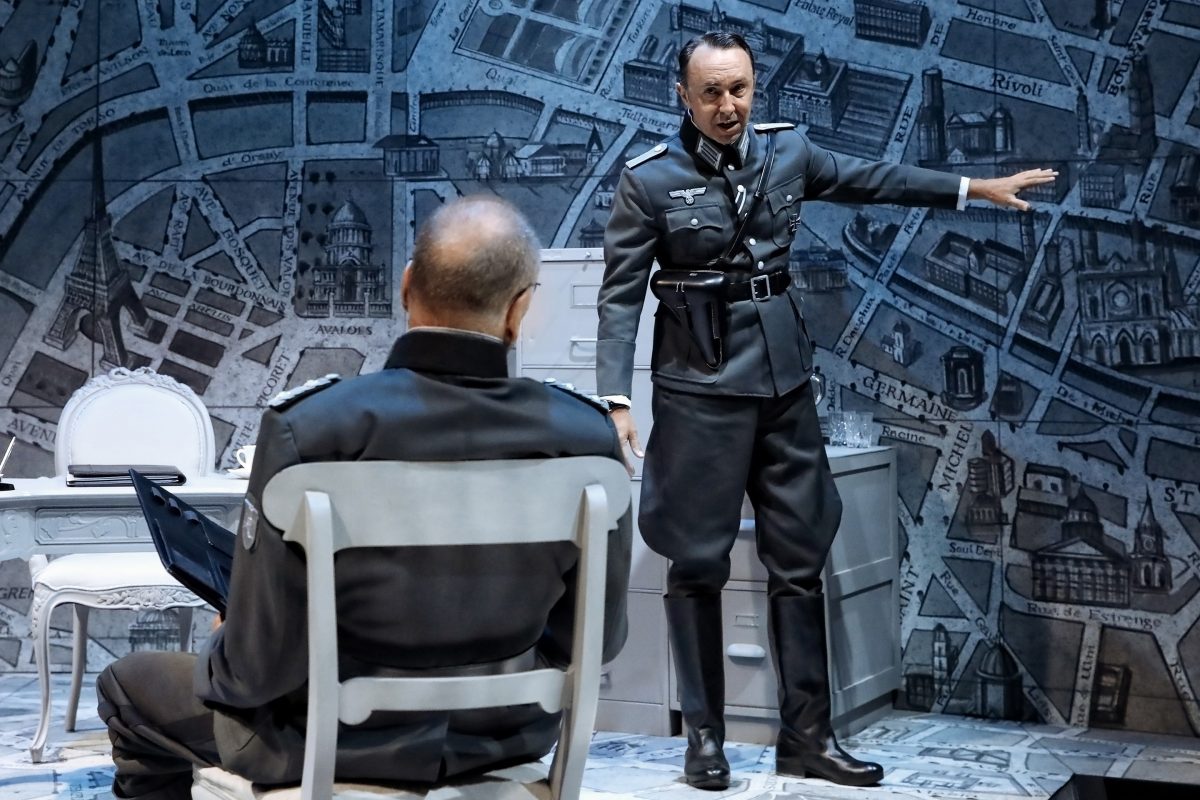 Two men in Second World War-era German military uniforms perform on stage; one sits facing away while the other stands gesturing, with a map-covered set as the backdrop, creating a tense, dramatic atmosphere.