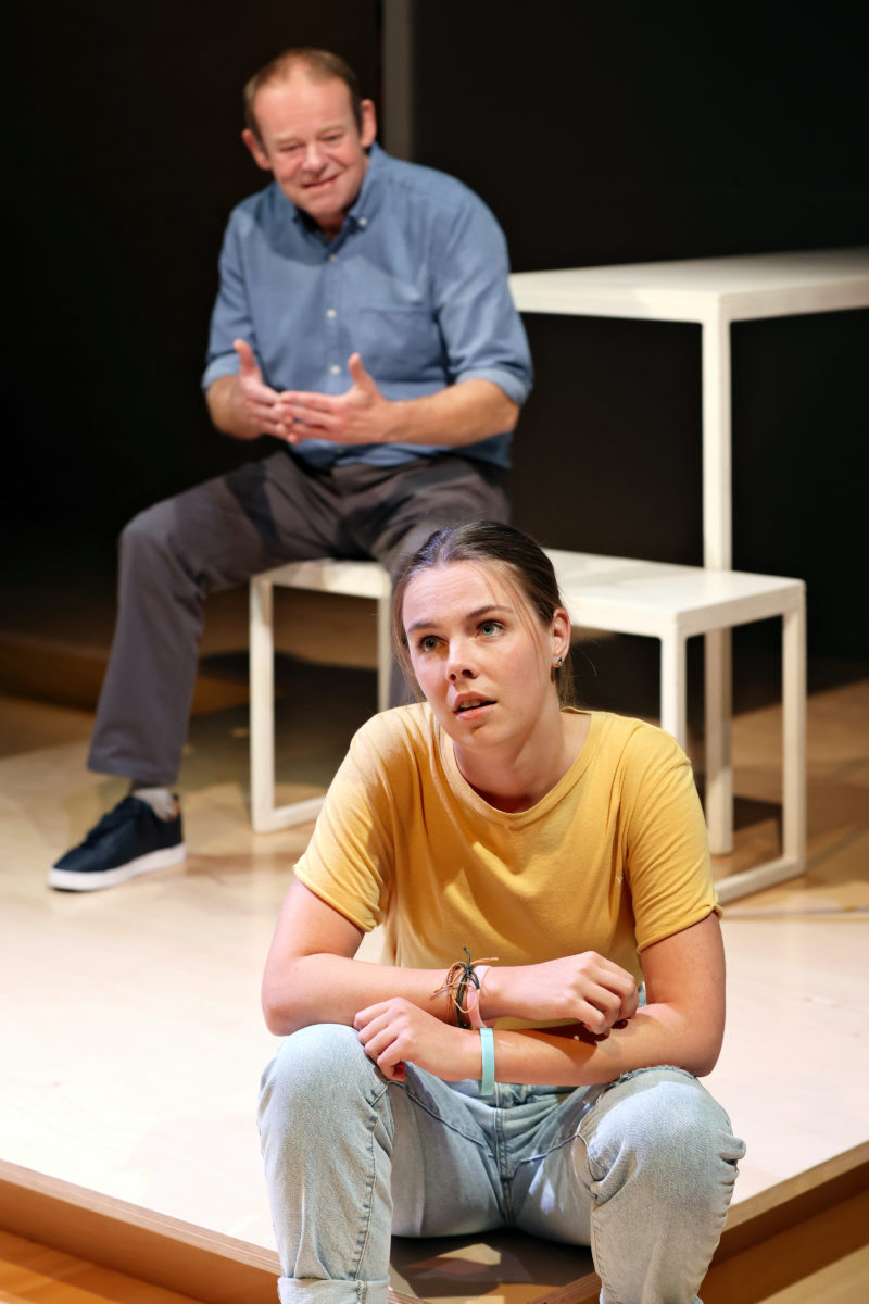 A young woman in a yellow shirt sits on the floor looking thoughtful, while an older man in a blue shirt sits behind her on a bench, gesturing with his hands as if speaking. Both are on a minimal stage.