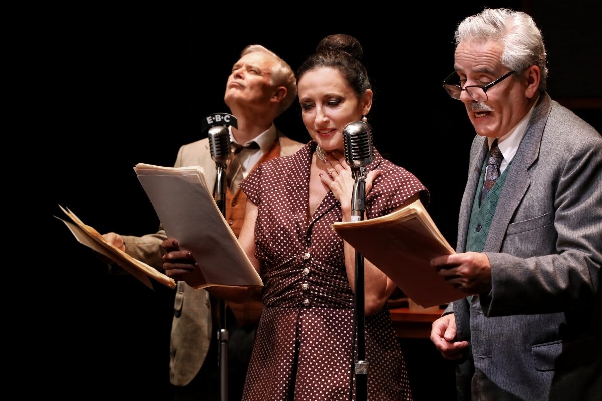Three actors in vintage attire stand at old-fashioned microphones, holding scripts and performing as if in a radio play. The woman in the centre smiles, while the two men on either side appear focused on their lines.