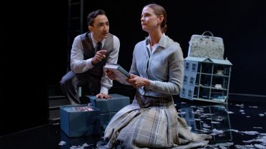A man and a woman in period clothing sit on a dark stage, surrounded by blue boxes, shredded paper, and a dolls' house. The woman kneels and holds a book, looking upward, while the man gestures, speaking to her.
