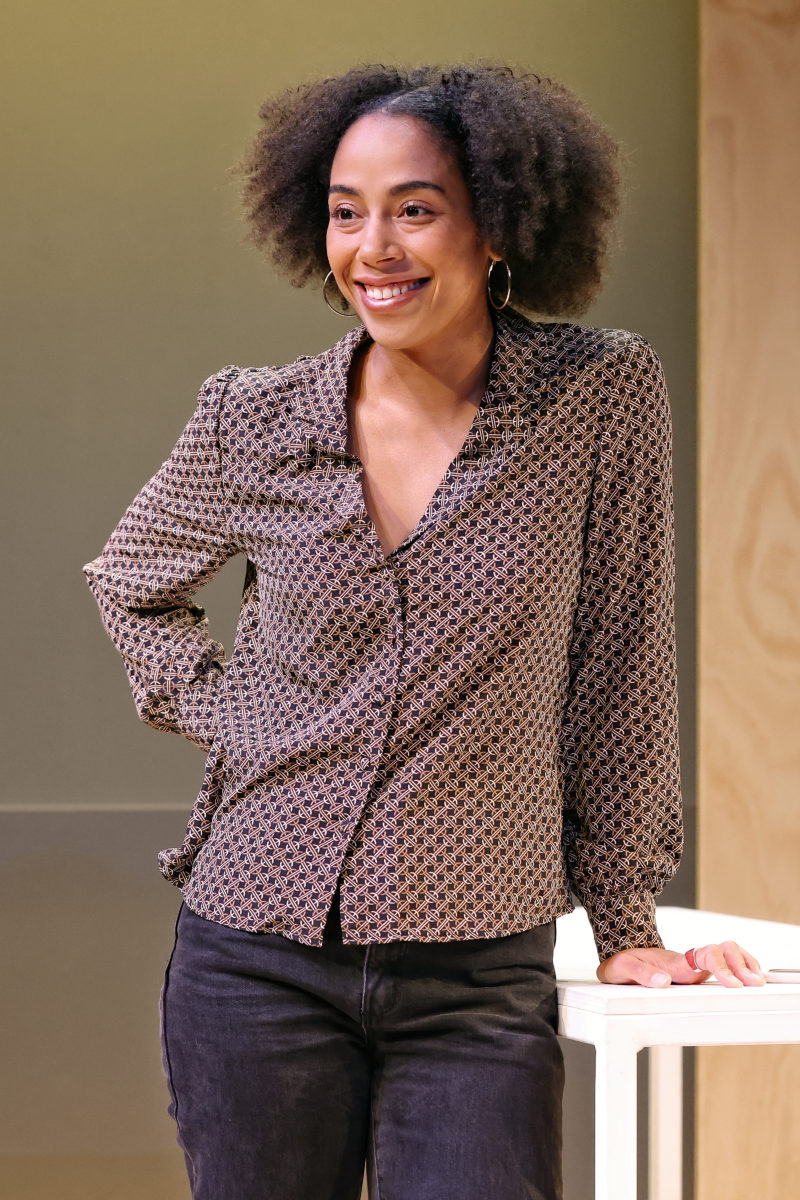 A woman with curly hair, wearing a patterned blouse and dark jeans, smiles whilst leaning on a white table with one hand. She stands in a brightly lit room with a neutral background.