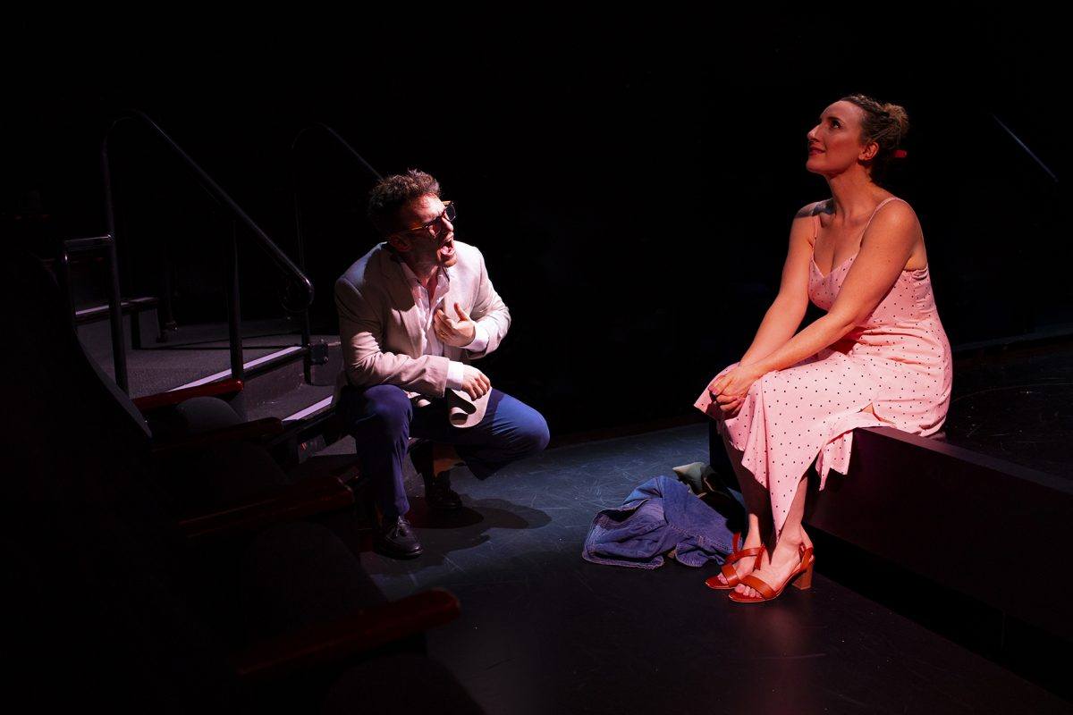 A man in a white jacket kneels and gestures towards a woman in a pink dress sitting on a stage edge, looking upwards and smiling under dramatic lighting in a dark theatre setting.