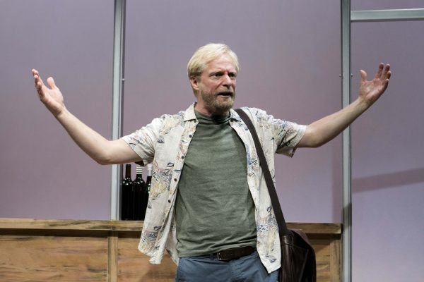 A man with blond hair and a beard, wearing a floral shirt and green T-shirt, stands with arms outstretched in front of a bar with wine bottles, appearing to speak passionately on stage.