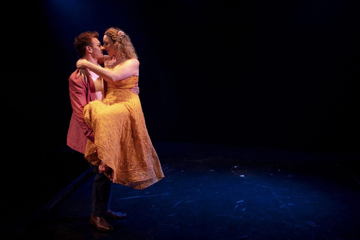 A man in a red jacket lifts a woman in a yellow dress under blue stage lighting. They gaze into each other’s eyes, standing on a dark stage with dramatic shadows.