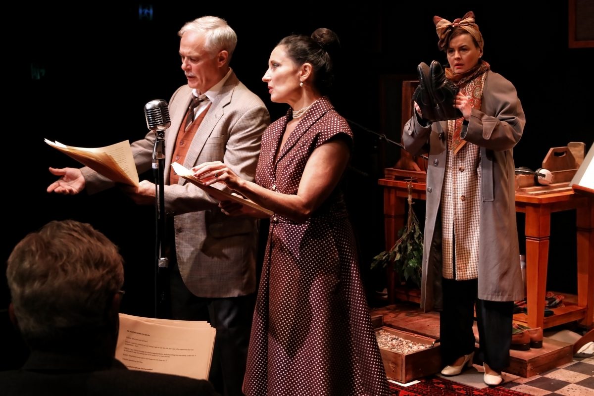 Three actors perform on stage in vintage attire, reading from scripts at a microphone. A woman in the background, wearing a coat and headscarf, holds up a pair of shoes and looks surprised. A seated person is visible in the foreground.