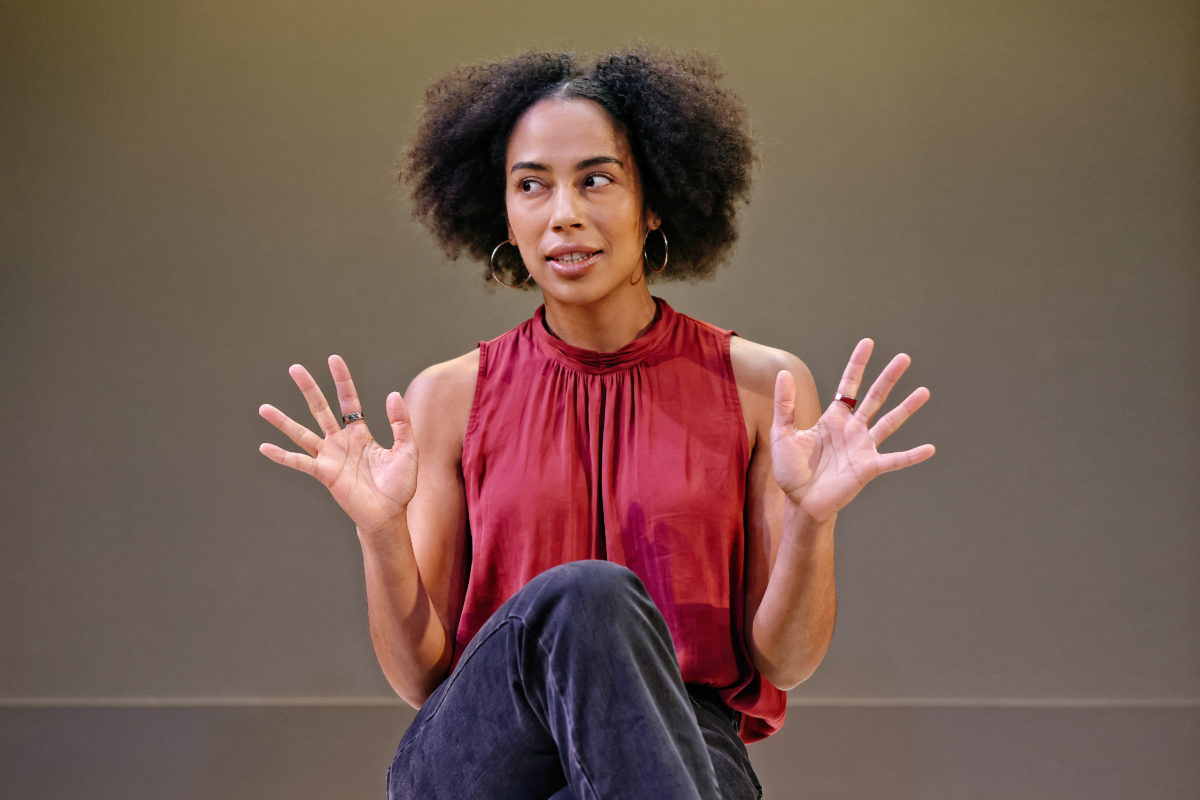 A woman with curly hair, wearing a sleeveless red top and dark trousers, sits and gestures with both hands raised, fingers spread apart, looking slightly to her left against a plain background.
