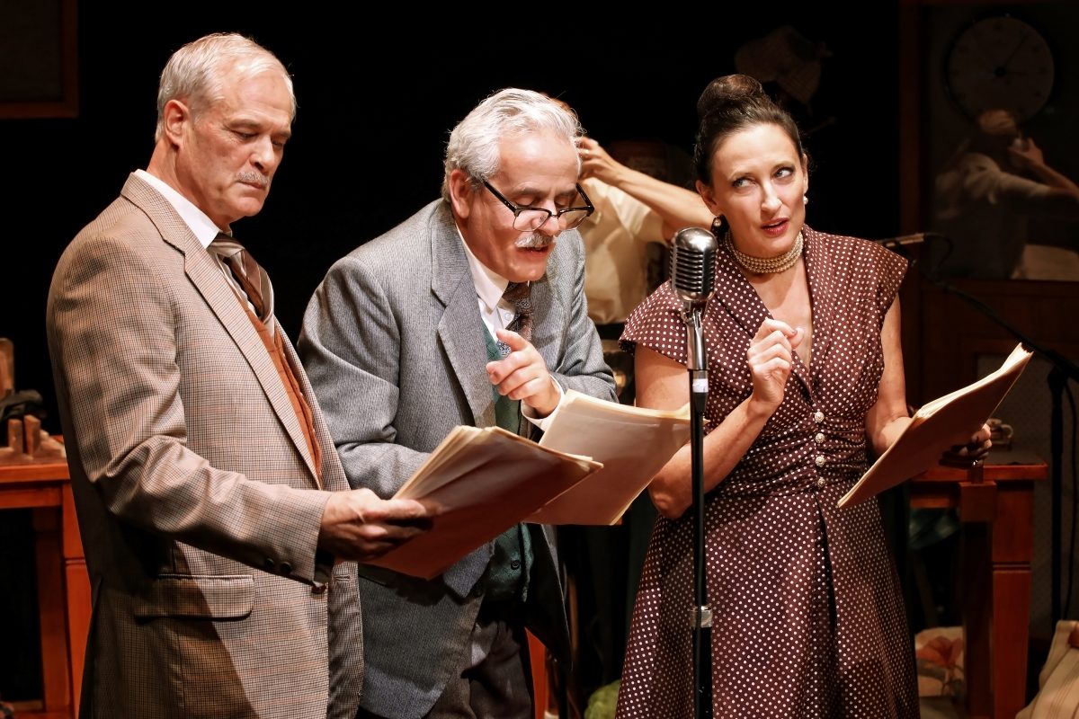 Three actors in vintage clothing stand at a microphone, holding scripts, and performing as if in a radio play. The background includes old-fashioned props and two partially visible figures.