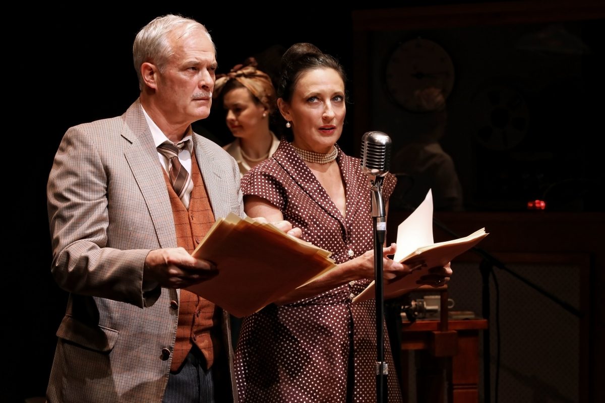 Two actors dressed in vintage clothing stand by a microphone, holding scripts, as a third person stands behind them. The scene appears to be set for an old-time radio show or theatrical performance.
