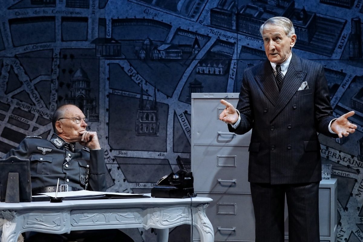 Two older men perform on stage; one in a military uniform sits at a desk, looking thoughtful, while the other in a pinstripe suit stands beside a filing cabinet, gesturing with open hands. A city map is in the background.