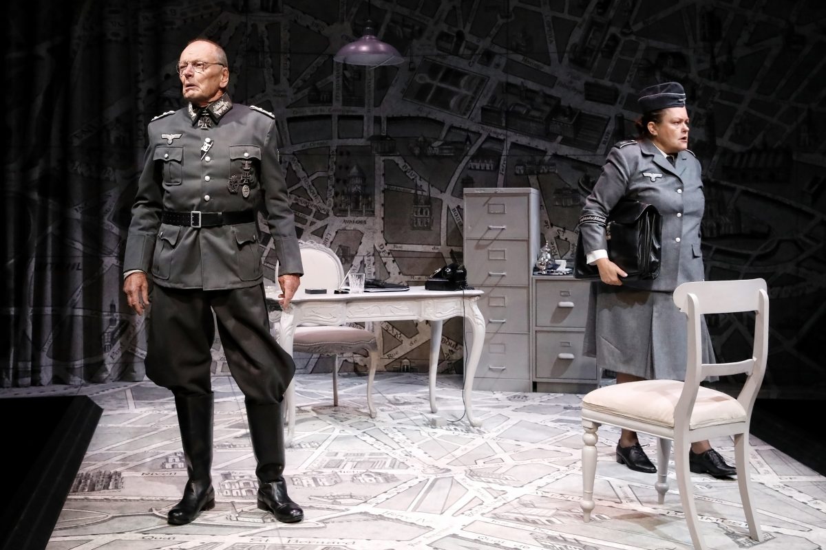 Two actors in Nazi military uniforms stand on a stage set resembling an office, with a desk, chair, and filing cabinet, against a backdrop of a large map. One stands confidently in front, while the other adjusts their belt near the desk.