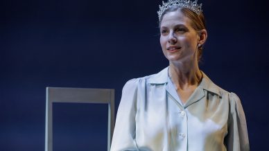 A woman wearing a light blue dress and a sparkling tiara sits in front of a simple chair, smiling gently against a dark background.