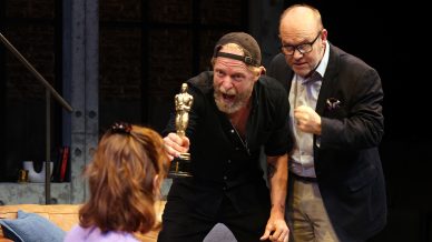 A bearded man in a backwards cap excitedly holds up an Oscar-like trophy towards a seated woman, whilst another man in glasses and a suit gestures emphatically beside him. They are on a stage set resembling a lounge.
