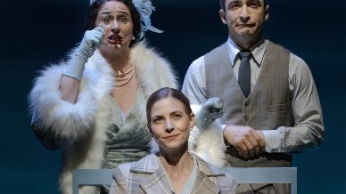 Three actors onstage: a woman in a green dress with white fur and gloves holds a handkerchief to her face, a man in a suit stands beside her, and a woman in a checked coat stands in front, all with dramatic expressions.
