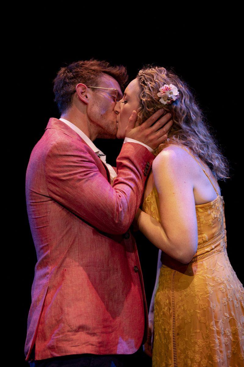 A man in a pink blazer and glasses kisses a woman in a yellow dress with a flower in her hair on stage, both standing close against a black background.
