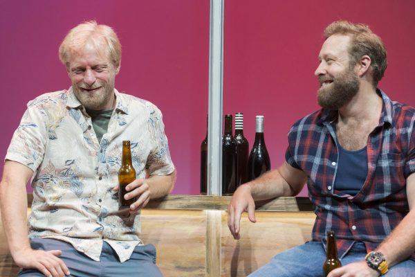 Two men sit on a wooden bench, smiling and holding beer bottles. The man on the left wears a patterned short-sleeve shirt, while the man on the right wears a checked sleeveless shirt. Several wine bottles are on the table behind them.