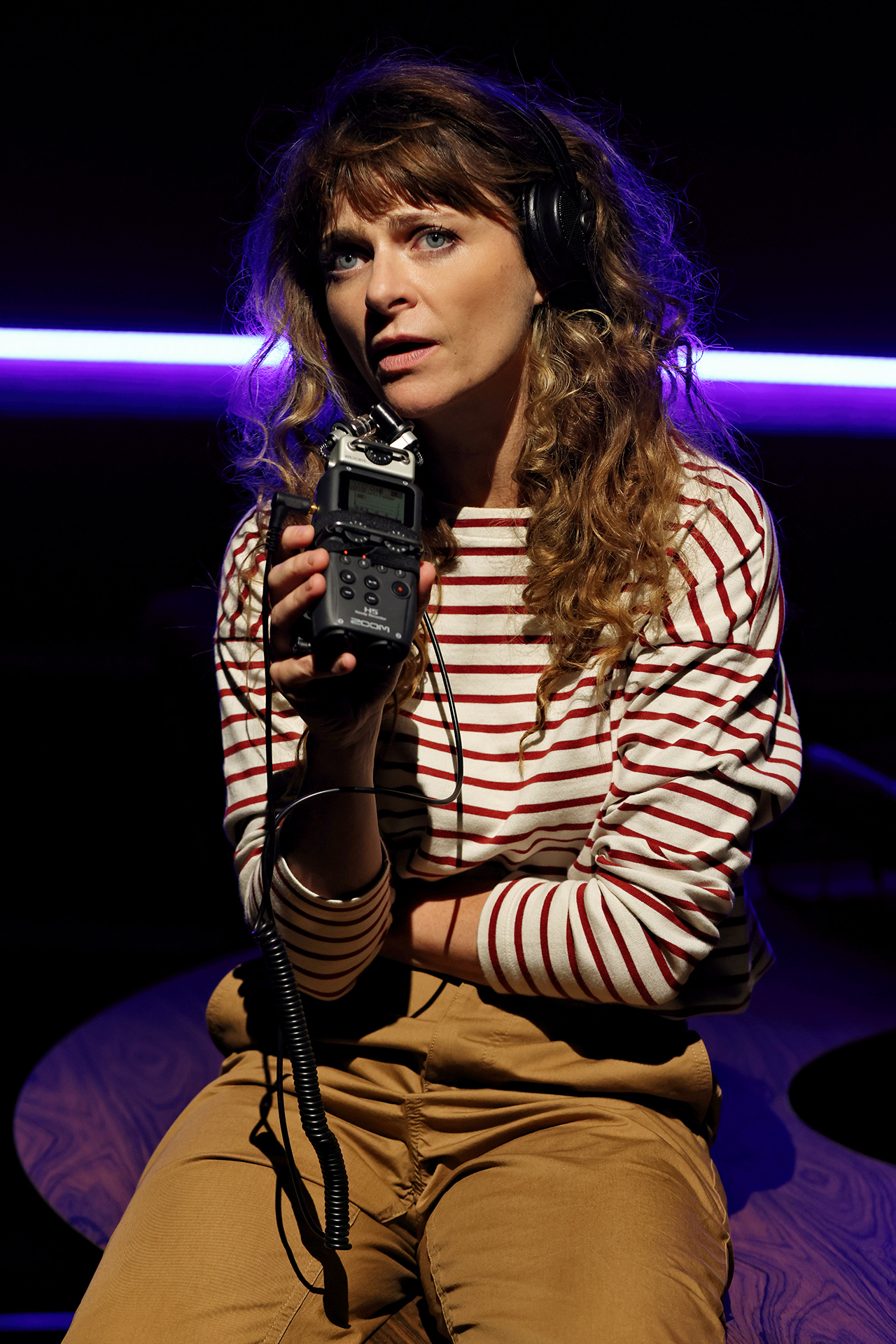 A woman with wavy hair, wearing headphones, a striped long-sleeved shirt, and tan trousers, holds a portable audio recorder and looks intently forward under dramatic lighting with a purple light strip in the background.