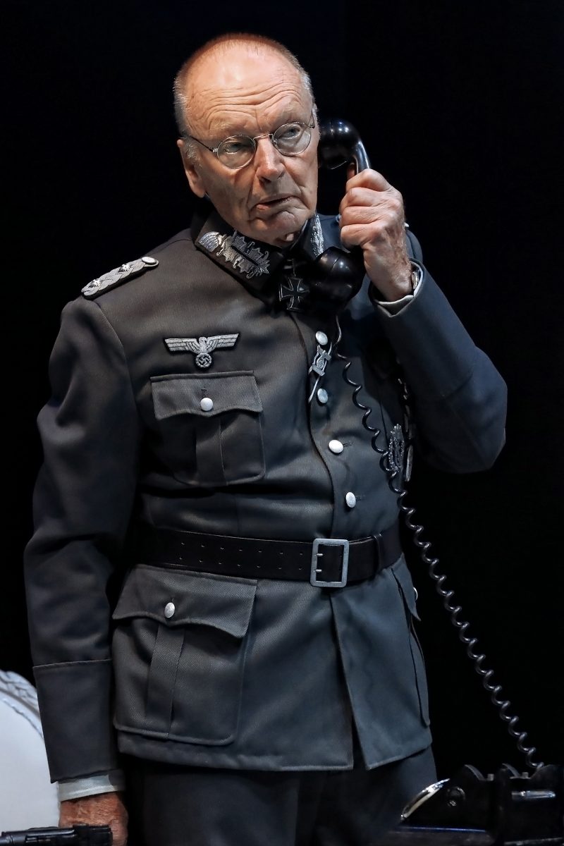 An older man in a Second World War-era German military uniform stands holding a black telephone handset to his ear, looking serious against a dark background.