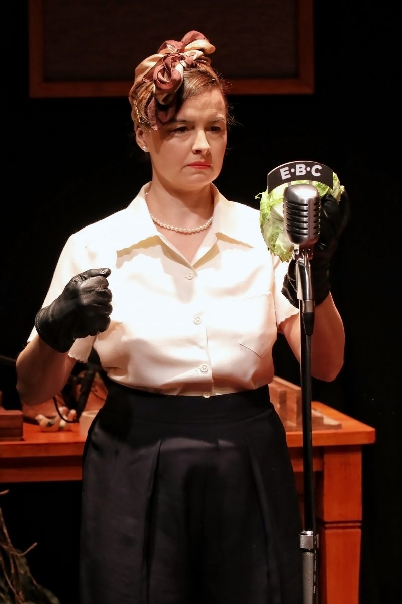 A woman wearing a headscarf, pearl necklace, white blouse, and black gloves stands in front of an old-fashioned microphone labelled EBC, holding a leafy cabbage in one hand, with a desk and objects in the background.