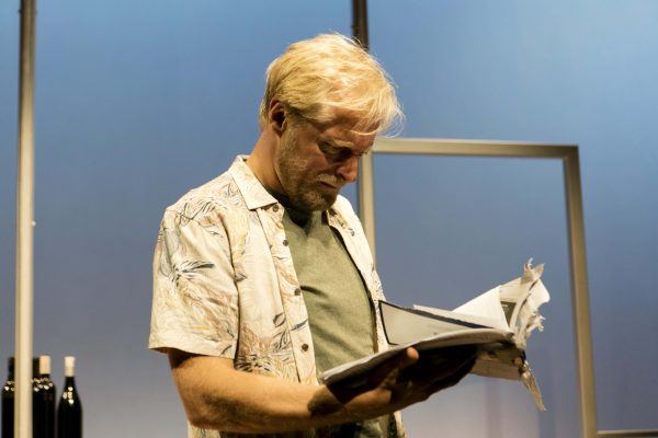 A man with blond hair and a beard, wearing a patterned short-sleeve shirt over a T-shirt, stands indoors and looks intently at an open, dishevelled book; three wine bottles are visible in the background.