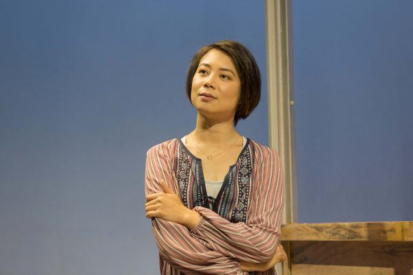 A woman with short dark hair stands with her arms folded, wearing a striped shirt with embroidered patterns, looking thoughtfully into the distance against a plain blue background.