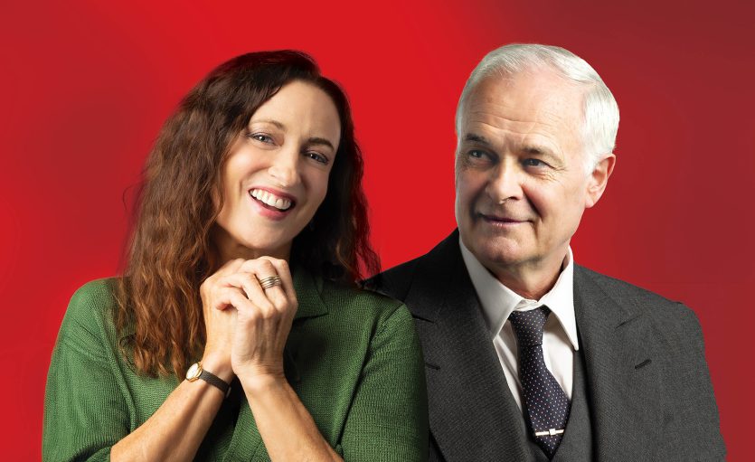 A smiling woman with long brown hair and a green top stands beside an older man in a suit and tie, both posed against a bright red background.