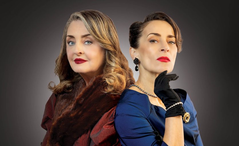 Two women pose back-to-back against a dark gradient background. One wears a fur stole and has wavy hair, while the other wears elegant gloves, a blue dress, and vintage earrings. Both have confident, serious expressions.