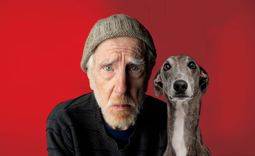 An elderly man wearing a knitted hat and jumper sits next to a greyhound against a bright red background, both looking directly at the camera with serious expressions.