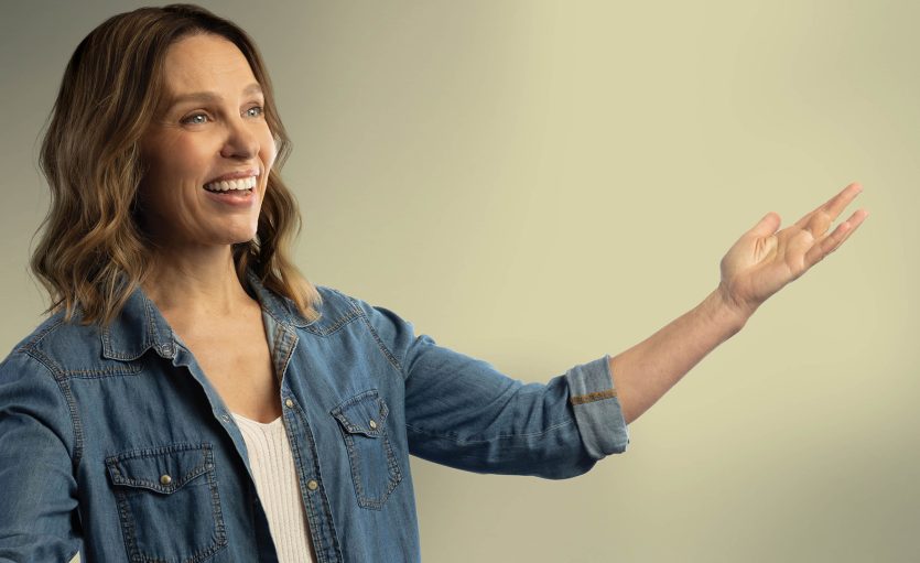 A smiling woman with wavy brown hair, wearing a denim shirt over a white top, gestures with one arm extended against a plain, light-coloured background.