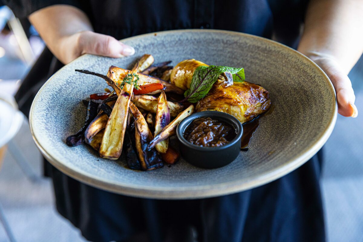 A person holds a plate with roast chicken, assorted roasted root vegetables, and a small dish of brown gravy, garnished with a sprig of fresh herbs.