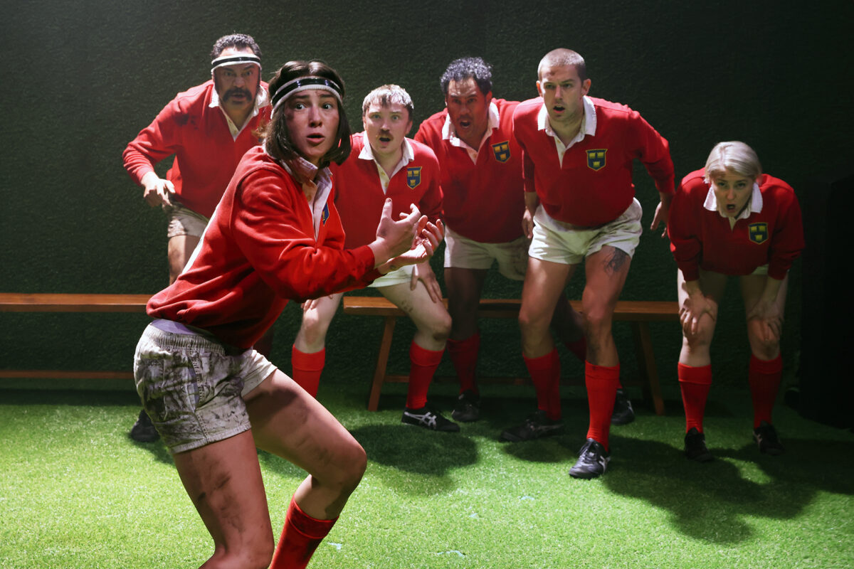 Six rugby players in red jerseys and red socks stand on artificial grass, looking alert and intense. One player in the foreground is holding an invisible ball, whilst the others crouch in anticipation.