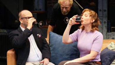 A man in glasses and a suit and a woman in a purple jumper sit on a sofa, both sipping wine, while another man with blond hair and a beard leans over behind them, watching closely.