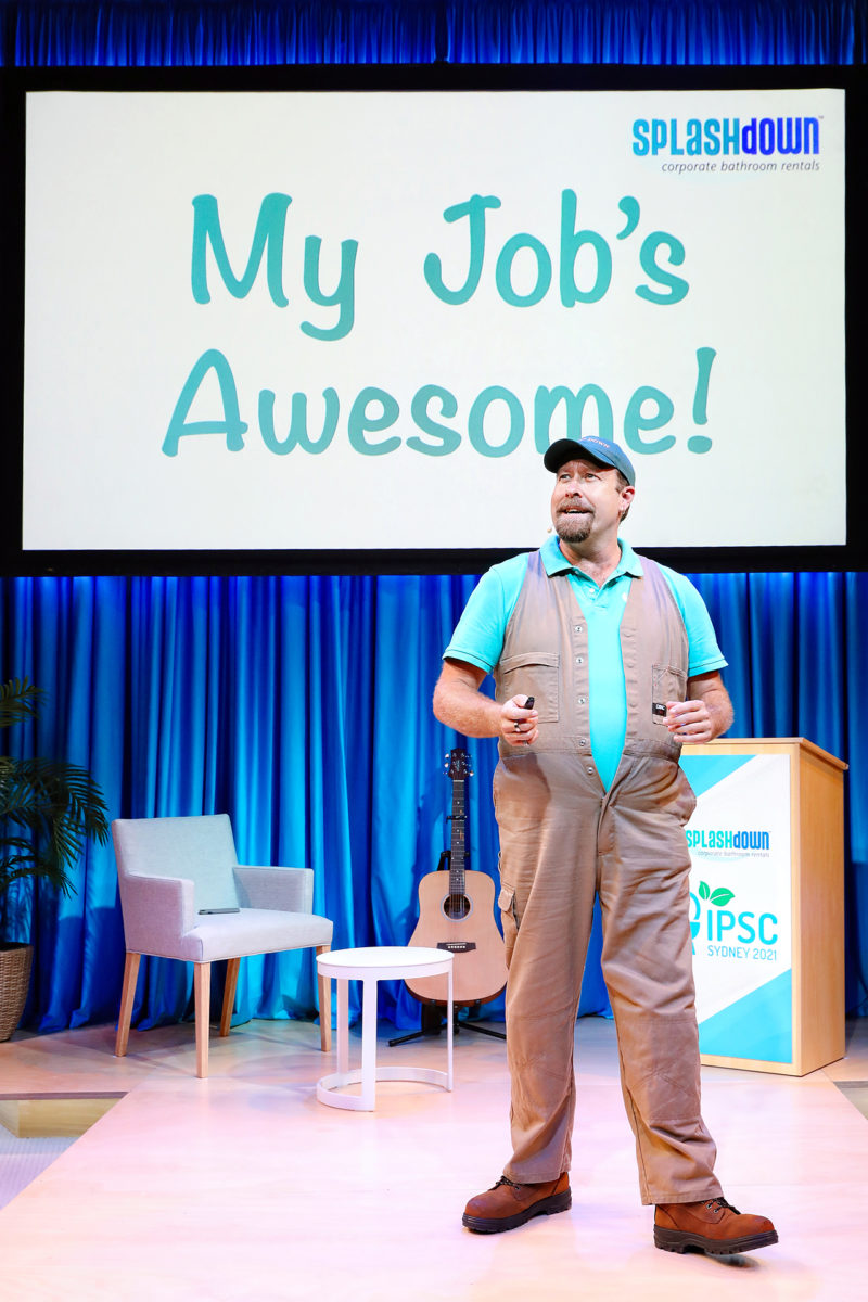 A man in khaki overalls and a teal shirt stands on stage with a presentation slide behind him that reads, “My Job’s Brilliant!” The stage has blue curtains, a chair, a guitar, and a small lectern.