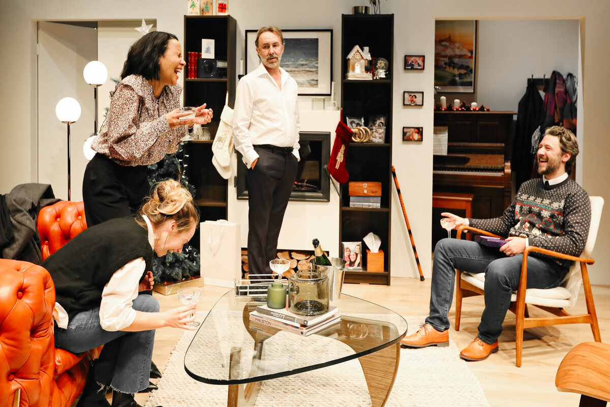 Four adults in a warmly lit sitting room laugh and converse. Two women are standing and crouching near a glass coffee table, while two men stand and sit comfortably, creating a lively, festive atmosphere.
