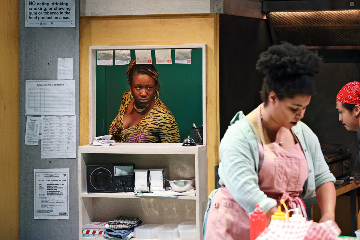 A woman in a leopard-print top looks through a serving hatch, while two people work in a kitchen. Signs, notes, and a radio are visible on the wall near the hatch. The kitchen appears busy and well-used.