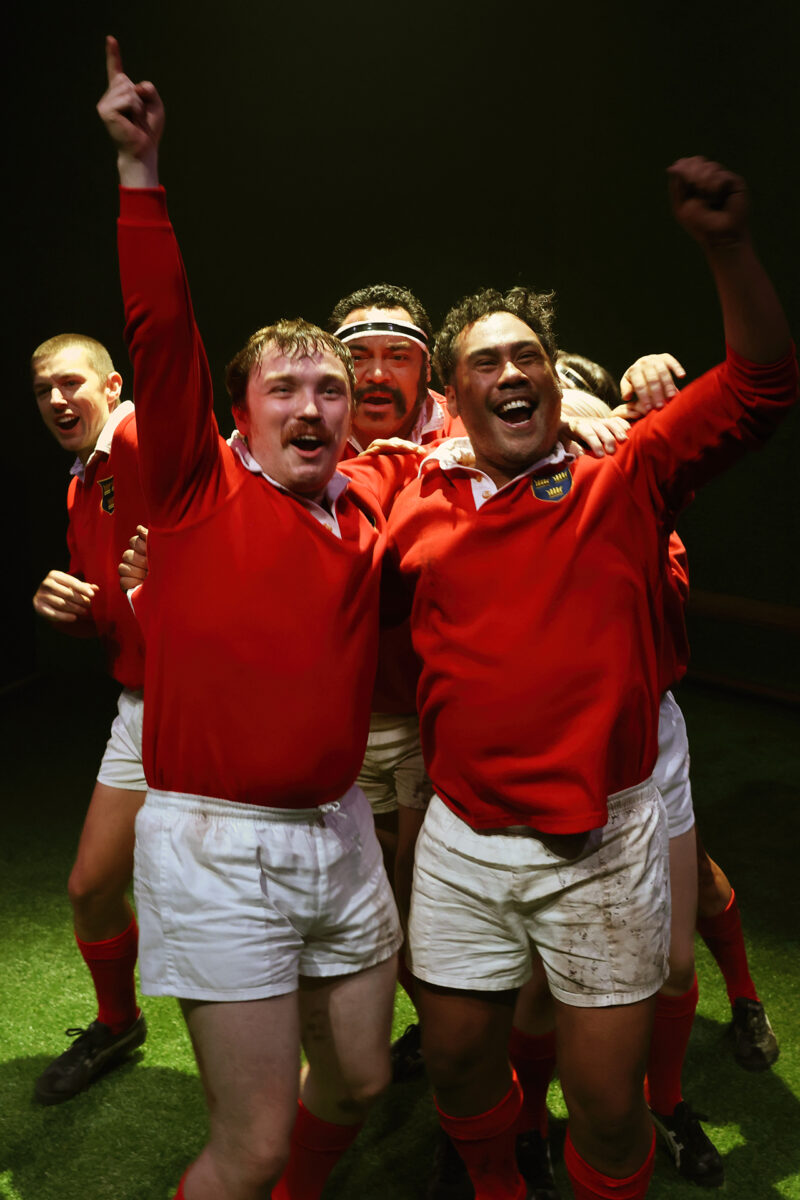 A group of rugby players in red jerseys and white shorts celebrate together on a pitch, smiling and raising their arms in victory.
