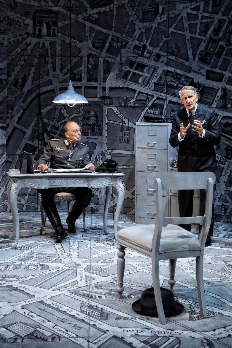 Two men in suits are on a stage set designed to look like a map; one sits at a desk with a lamp, typewriter, and telephone, while the other stands and gestures. A chair and hat are in the foreground.