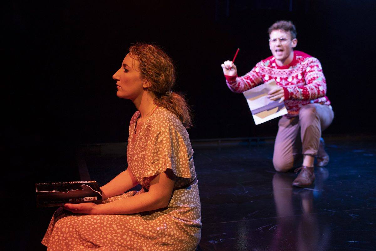 A woman in a light patterned dress sits on the floor holding two books, looking thoughtful, while a man in glasses and a red jumper kneels behind her, holding papers and a red pen, appearing to speak passionately.
